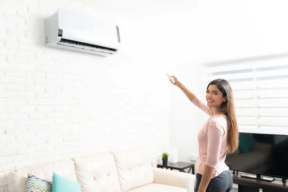 Woman smiling and using a remote control to operate a wall-mounted air conditioner in a bright living room.