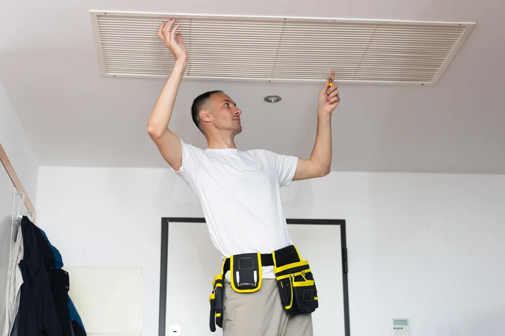 Technician inspecting a ceiling air vent with a screwdriver, wearing a tool belt and standing on a step ladder.