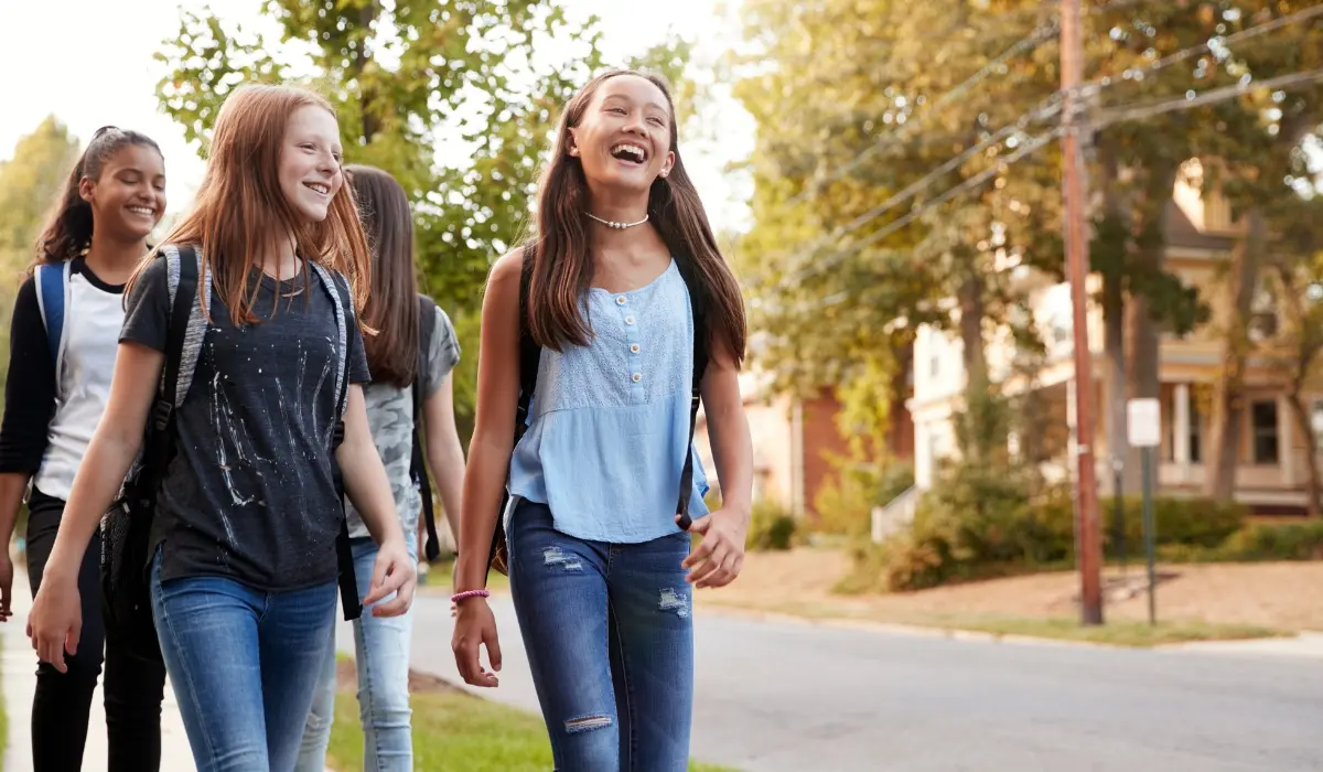 A group of young girls walk together outdoors, laughing and chatting. They seem to be enjoying each other's company, with a neighborhood street, trees, and residential houses in the background.