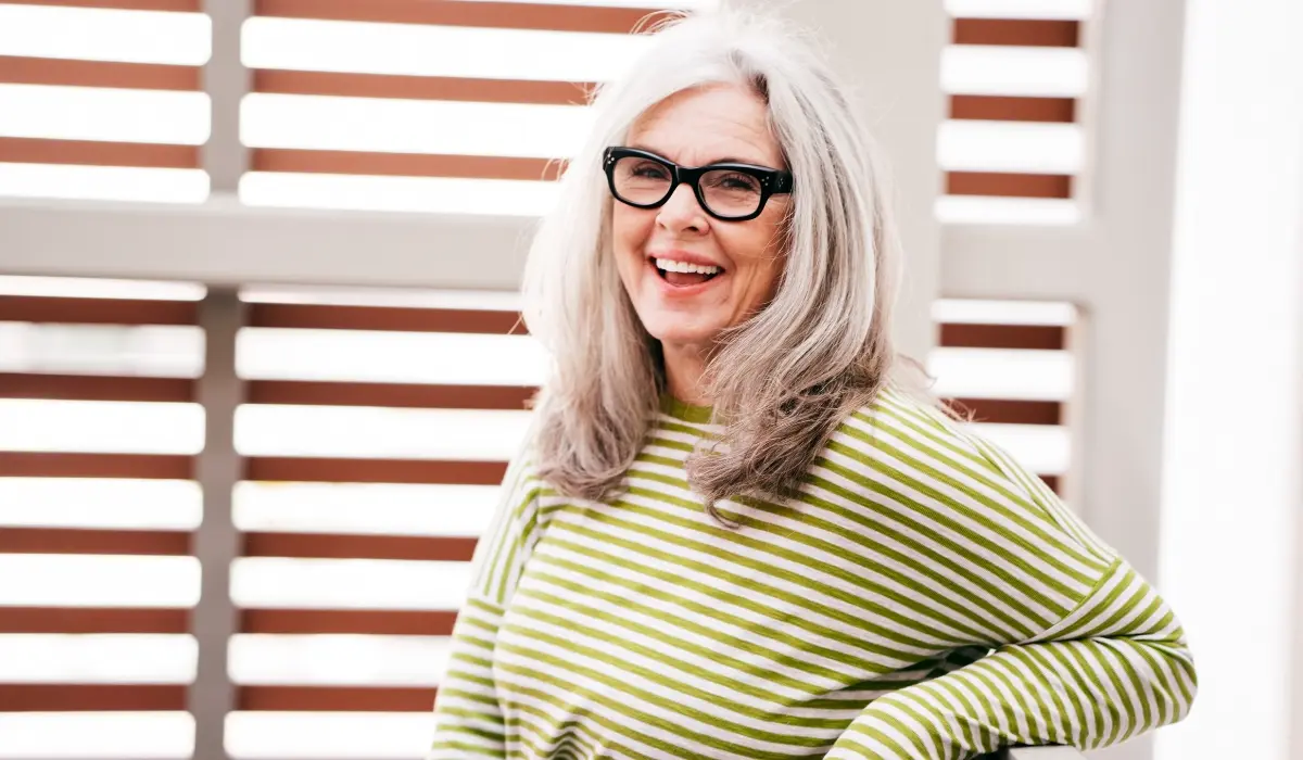 A smiling older woman with long gray hair, wearing black glasses and a green and white striped sweater. She is posing in front of a wooden slatted backdrop.