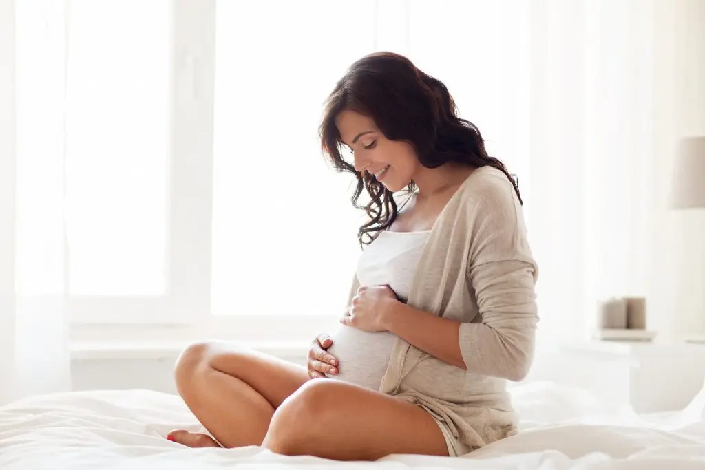 A smiling, pregnant woman sitting comfortably on a bed, gently holding her baby bump while looking down at it. She is wearing a light, cozy outfit and the soft, natural light coming through the window highlights the peaceful moment.