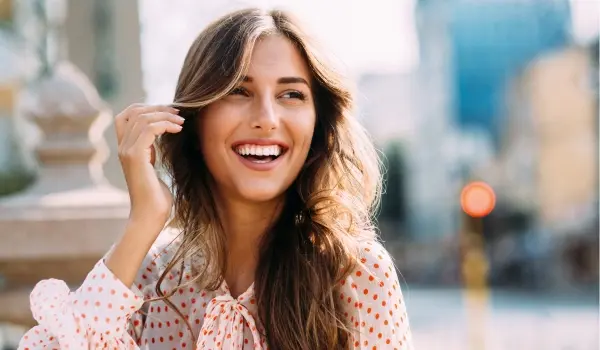A woman with long, wavy hair, smiling and playfully touching her hair while wearing a polka dot blouse in a sunny urban setting.