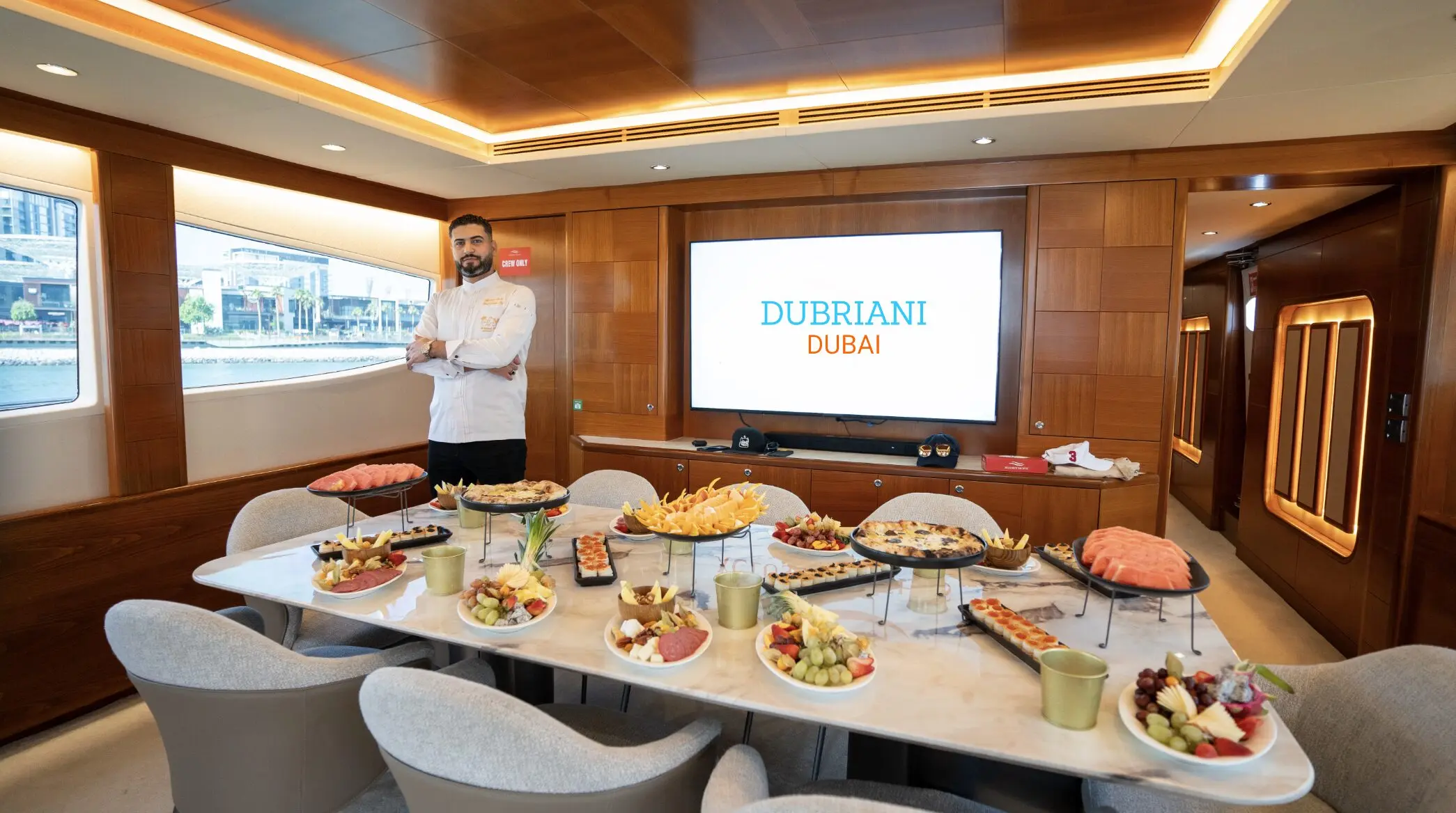 Chef standing beside a long buffet of fresh platters laid out in a wood-panelled yacht dining salon