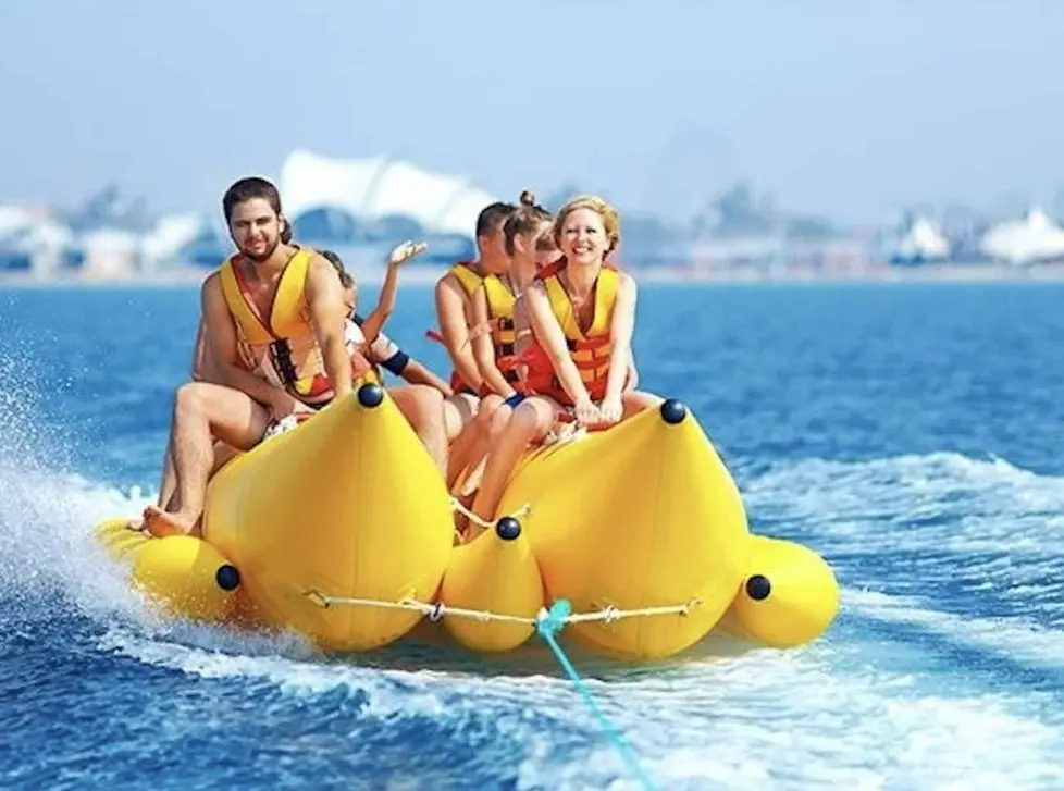 Group of five in orange life jackets laughing on a yellow banana boat towed across blue water