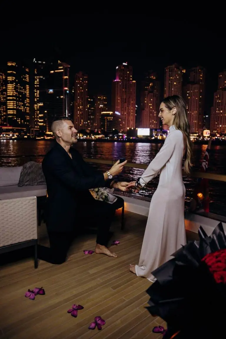 Man proposing on one knee to a woman on a yacht at night with a city skyline illuminated in the background.
