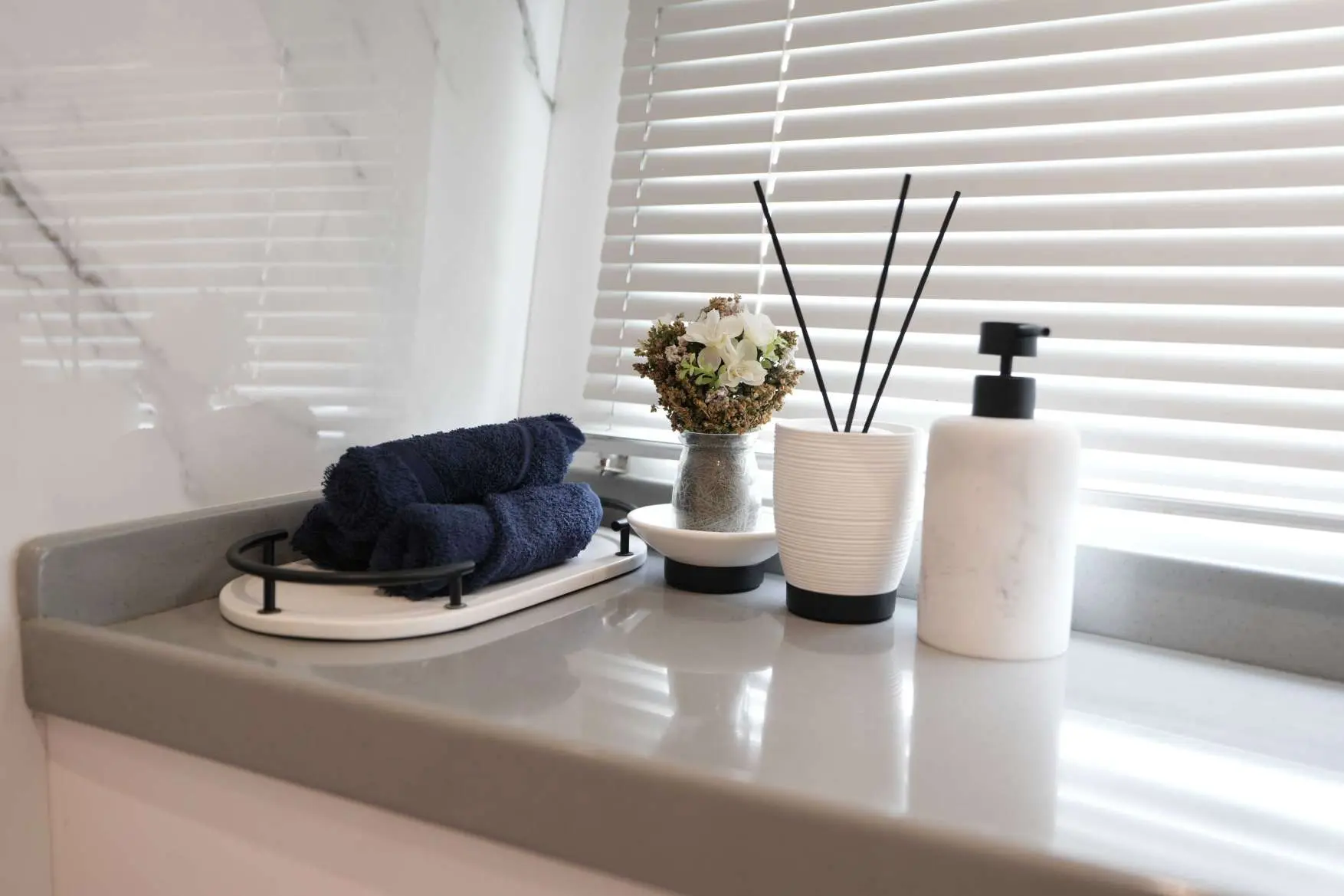 Luxury bathroom countertop on Belle 75 yacht with folded navy towels, marble soap dispenser, reed diffuser, and decorative flowers beside window blinds.