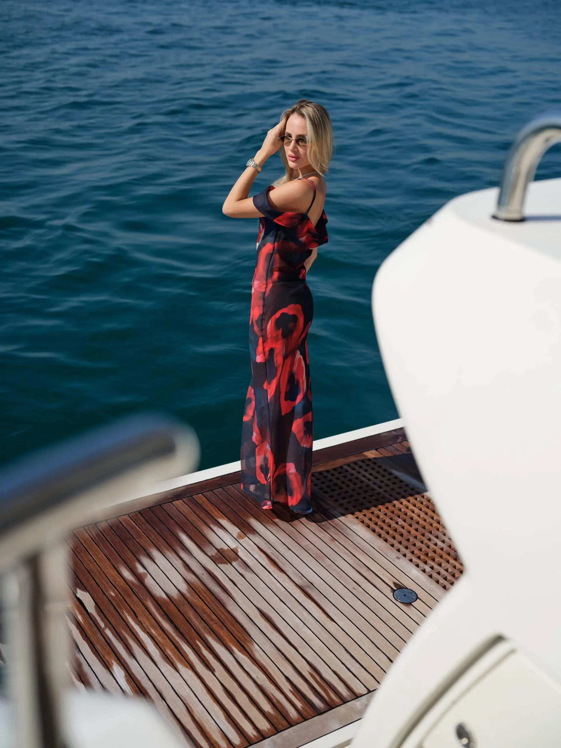 Guest in a long printed dress standing on a yacht's teak deck beside the white hull over blue water