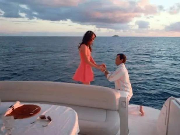 Man proposing on one knee to a woman in a pink dress aboard a yacht at sunset, surrounded by open sea.