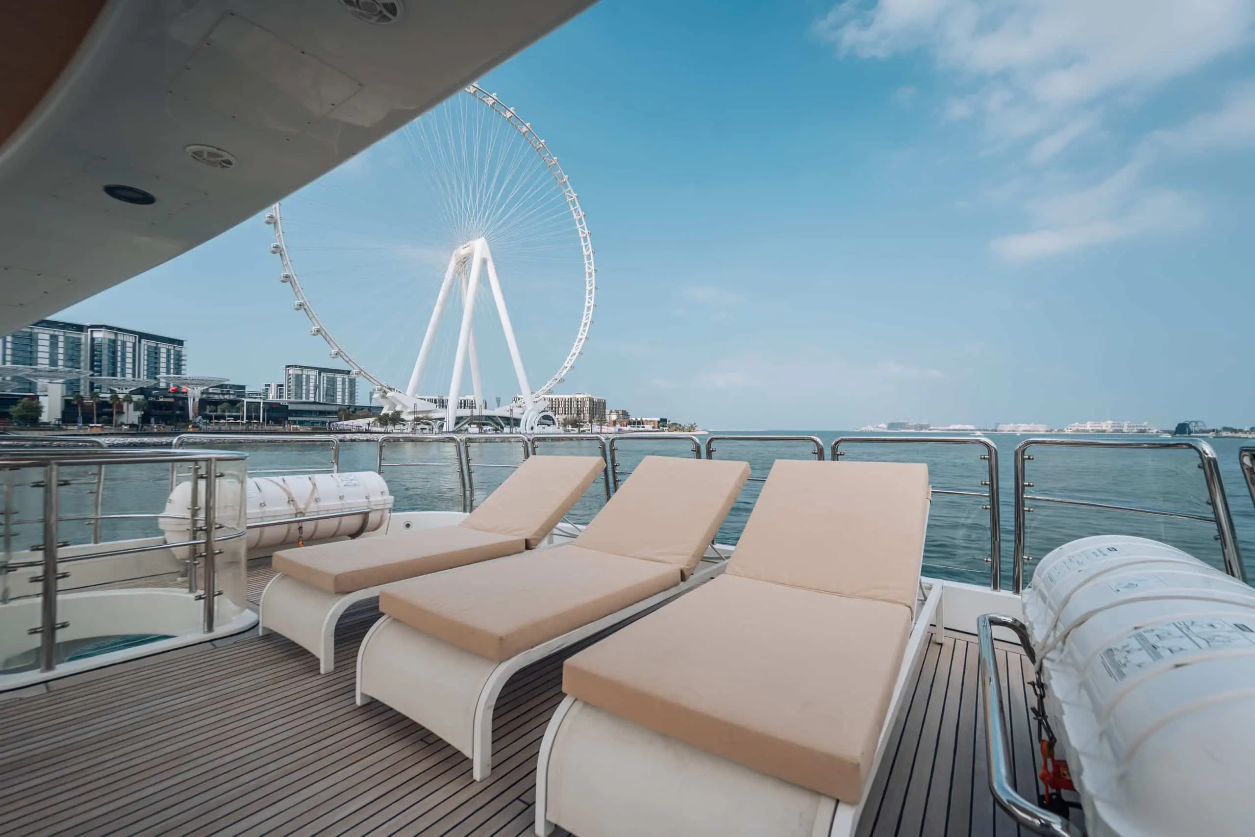Three beige sun loungers on a yacht deck overlooking the water, with Ain Dubai visible against a clear blue sky.