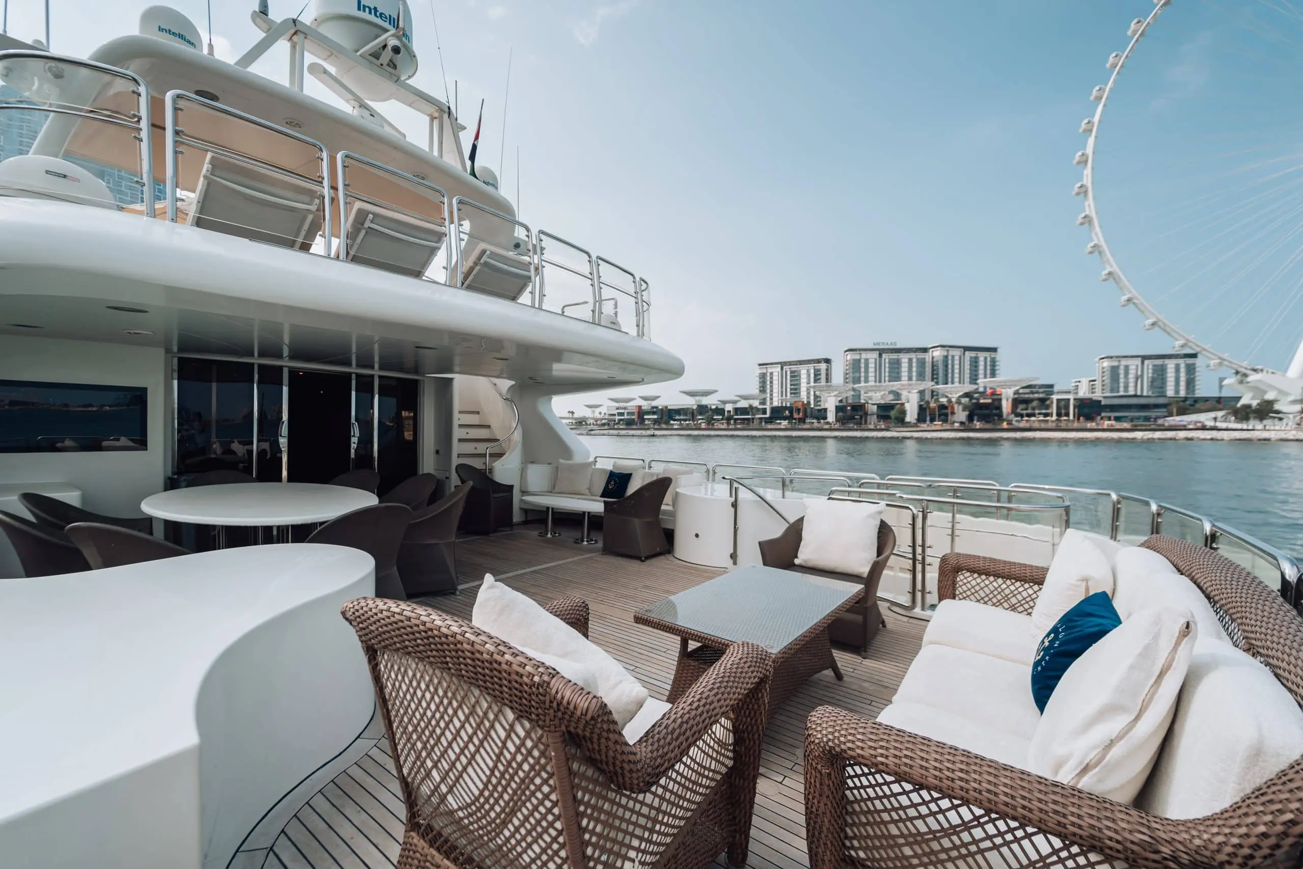 Shaded seating and dining area on the aft deck of a luxury yacht, with wicker lounge furniture, round tables, and a waterfront skyline with a large observation wheel in the background.