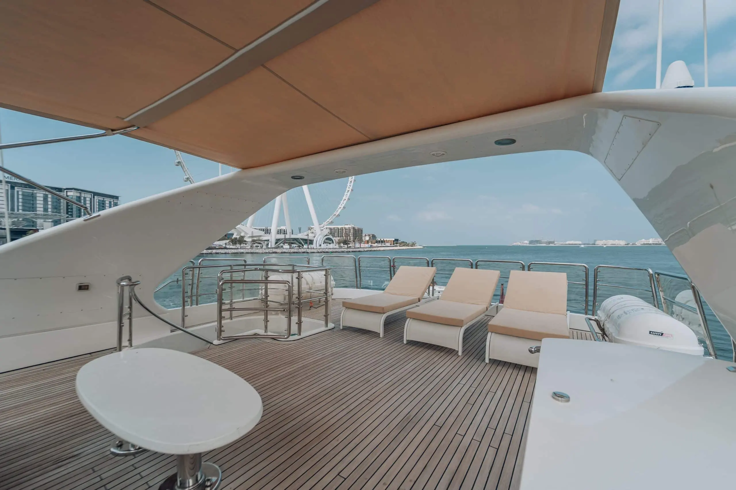 Luxury yacht deck with beige sun loungers under a canopy, overlooking turquoise water and Ain Dubai in the distance.