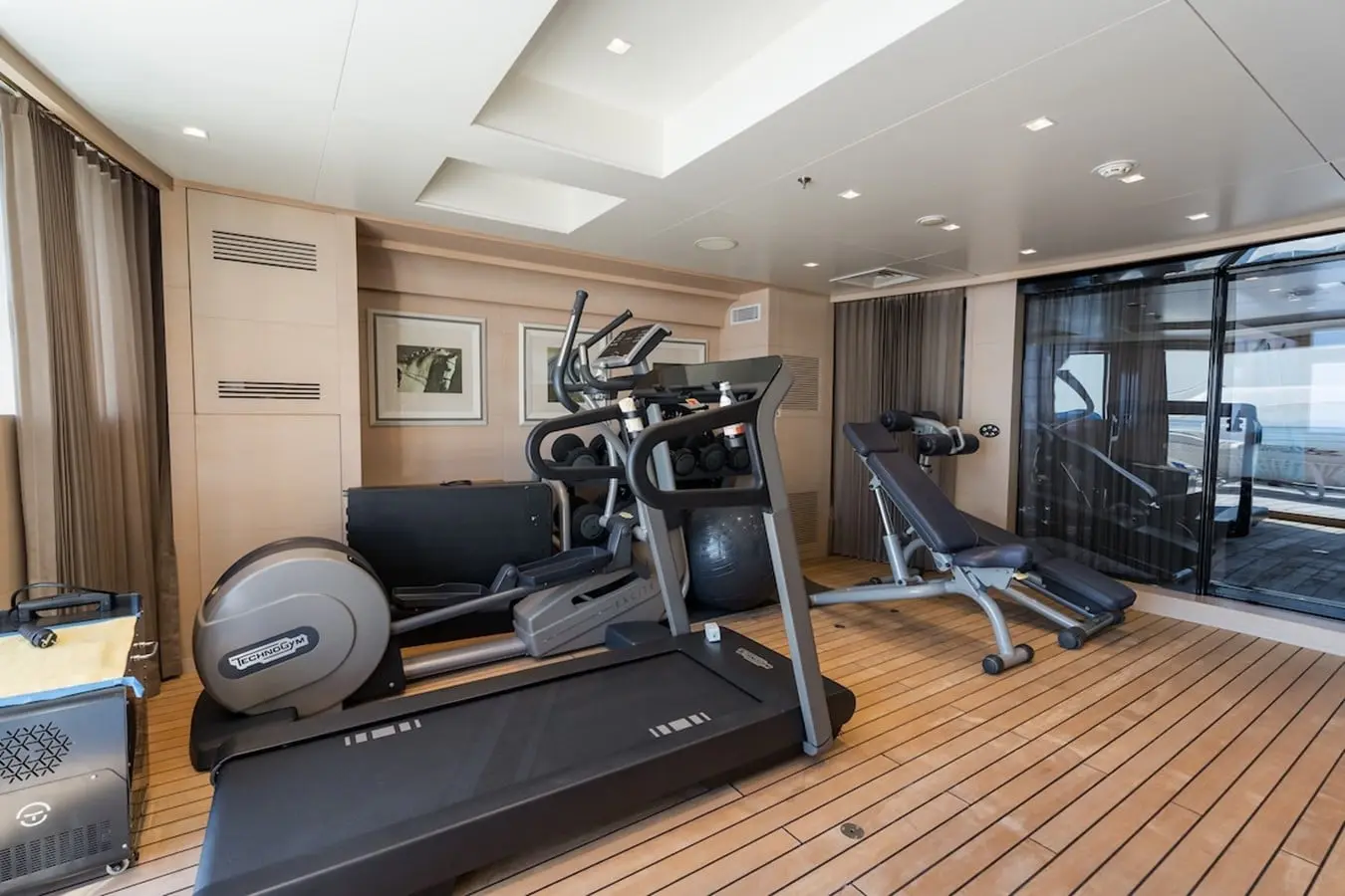 Yacht gym on teak decking with treadmill, cross-trainer, weight bench and dumbbells by glass doors