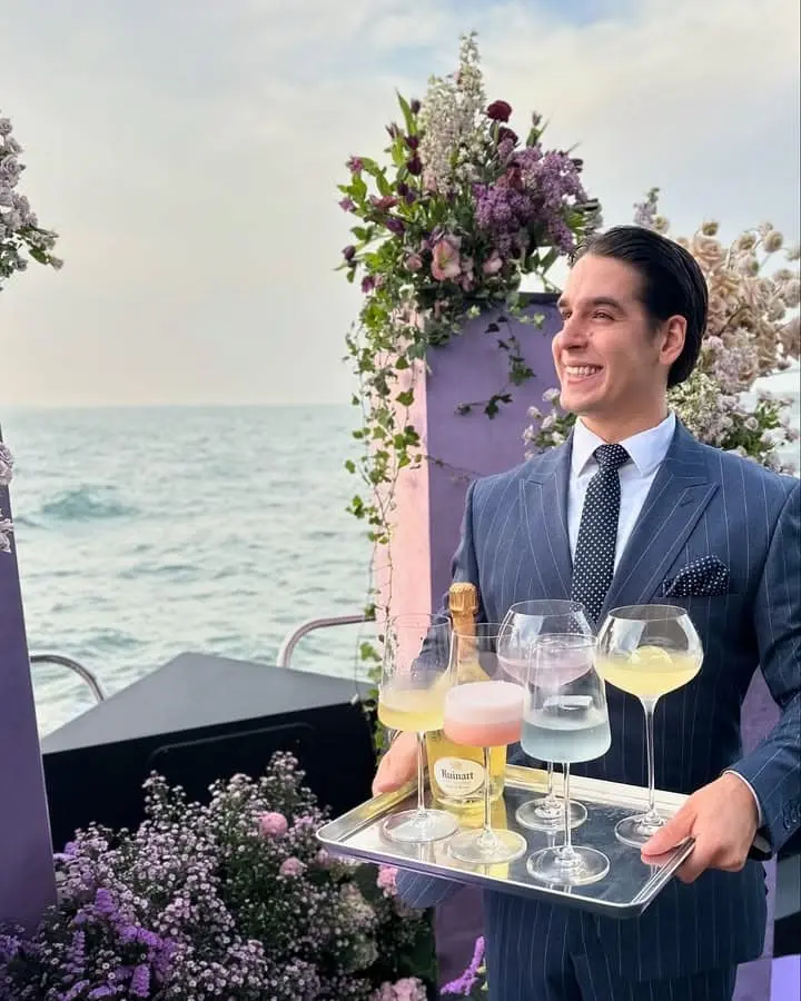 Server in a pinstripe suit carrying a tray of champagne glasses beside floral arrangements on deck at sea