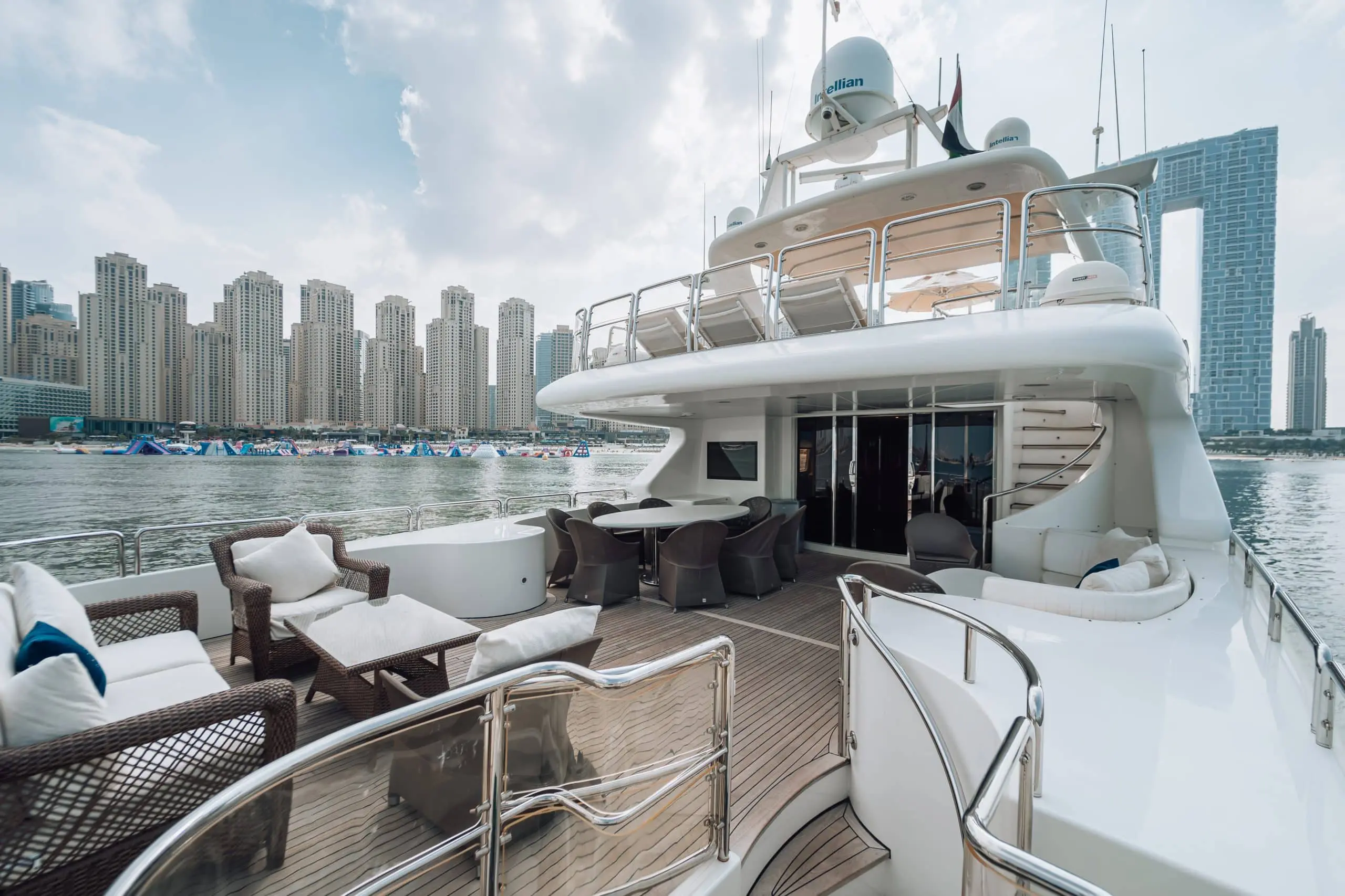 Spacious aft deck of a luxury yacht with wicker lounge seating and dining tables, overlooking calm water and a dense modern waterfront skyline with high-rise towers in the background.