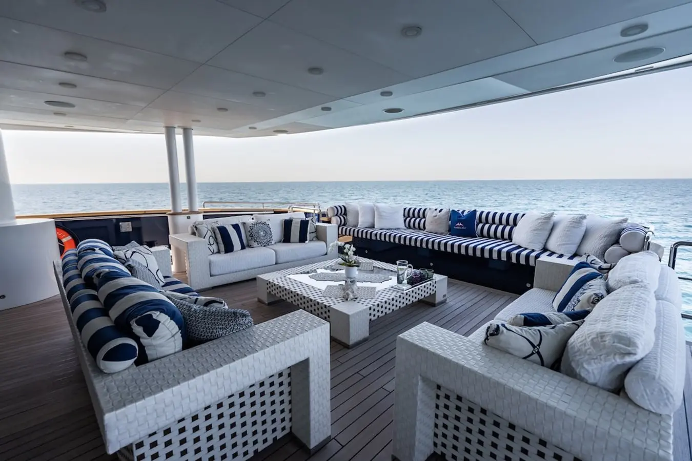 Covered aft deck lounge with white sofas, navy striped cushions and an open sea view on three sides