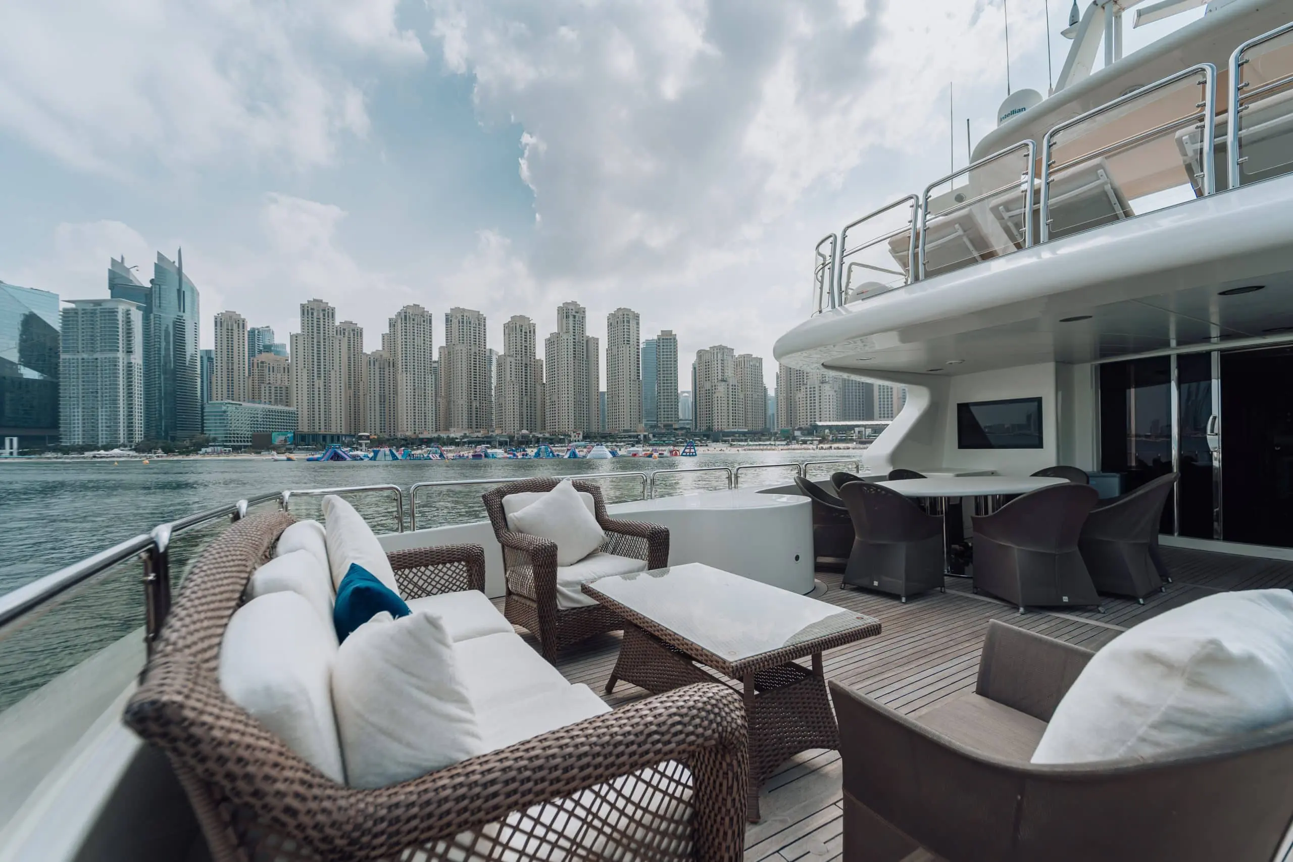 Wicker lounge seating and a low table on the open aft deck of a luxury yacht, overlooking calm water and a dense skyline of modern high-rise buildings across the marina.