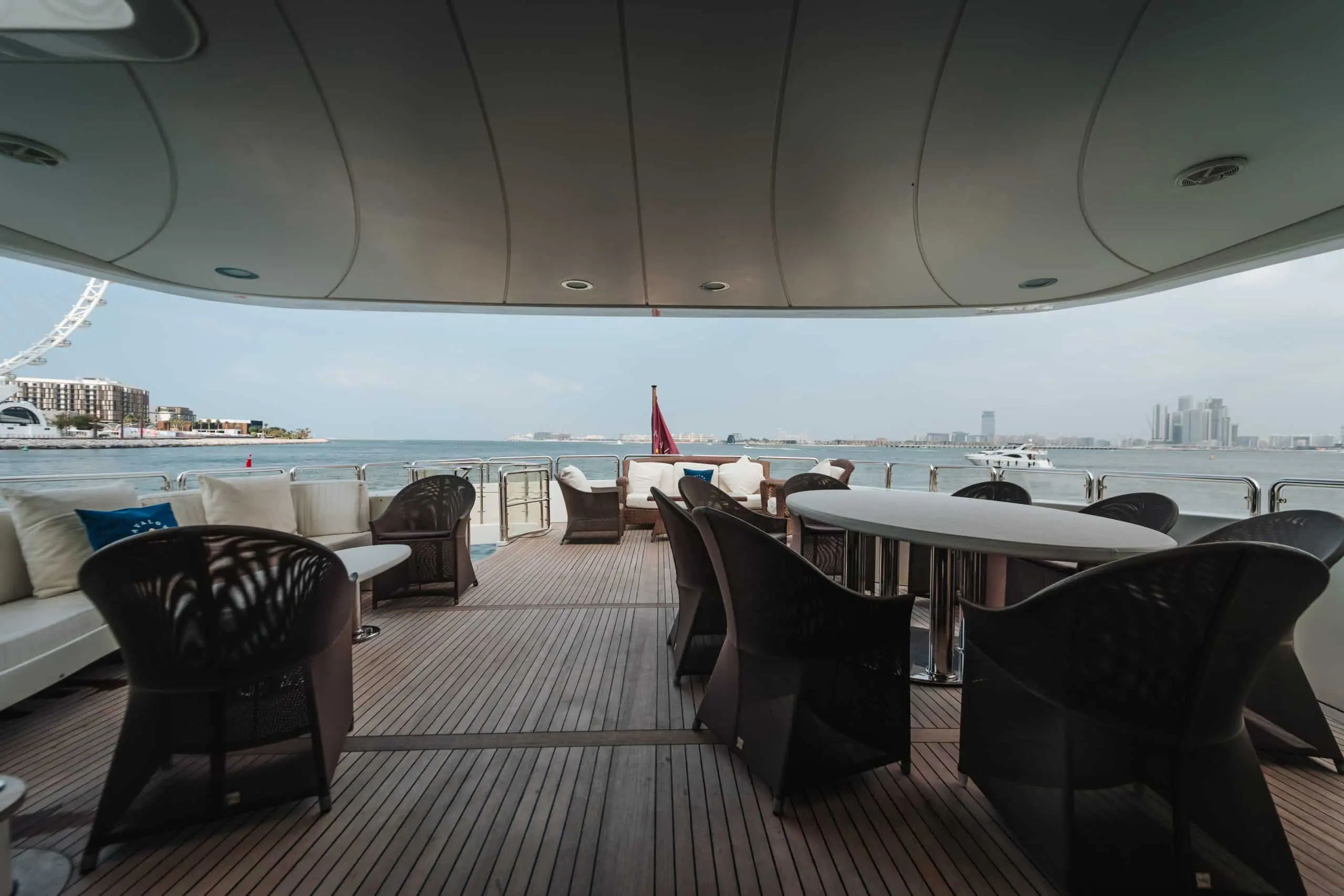 Outdoor seating area on the shaded upper deck of a luxury yacht, with tables and chairs overlooking calm water, a marina, and a modern city skyline in the distance.
