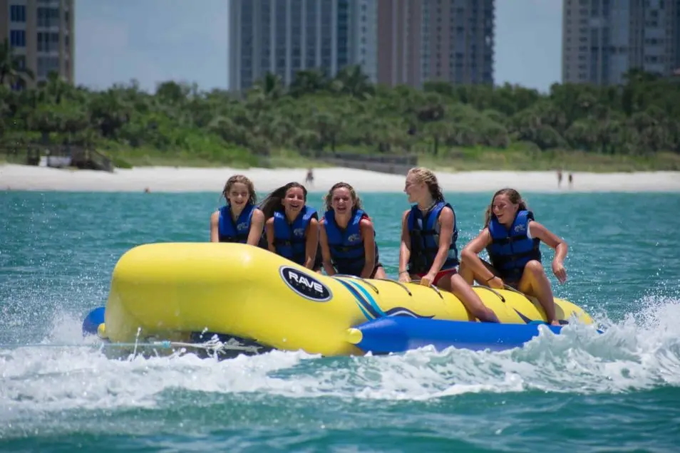 Five riders in blue life jackets on a yellow inflatable banana boat being towed near a sandy beach