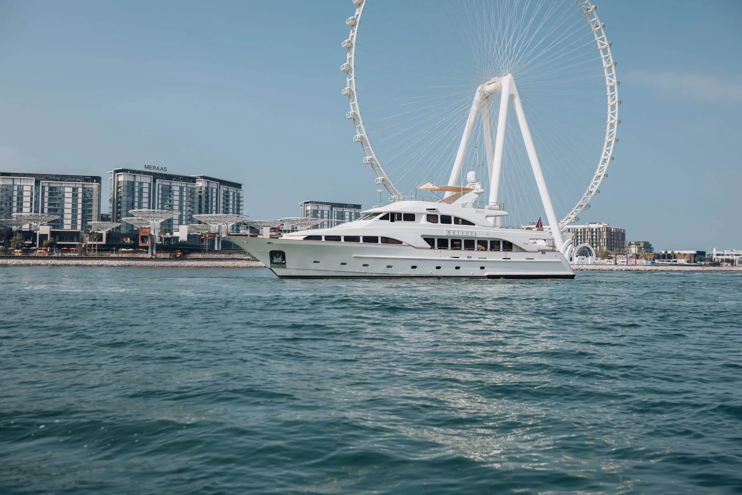 White luxury yacht sailing across turquoise waters with Ain Dubai and modern waterfront buildings in Dubai under a clear blue sky.