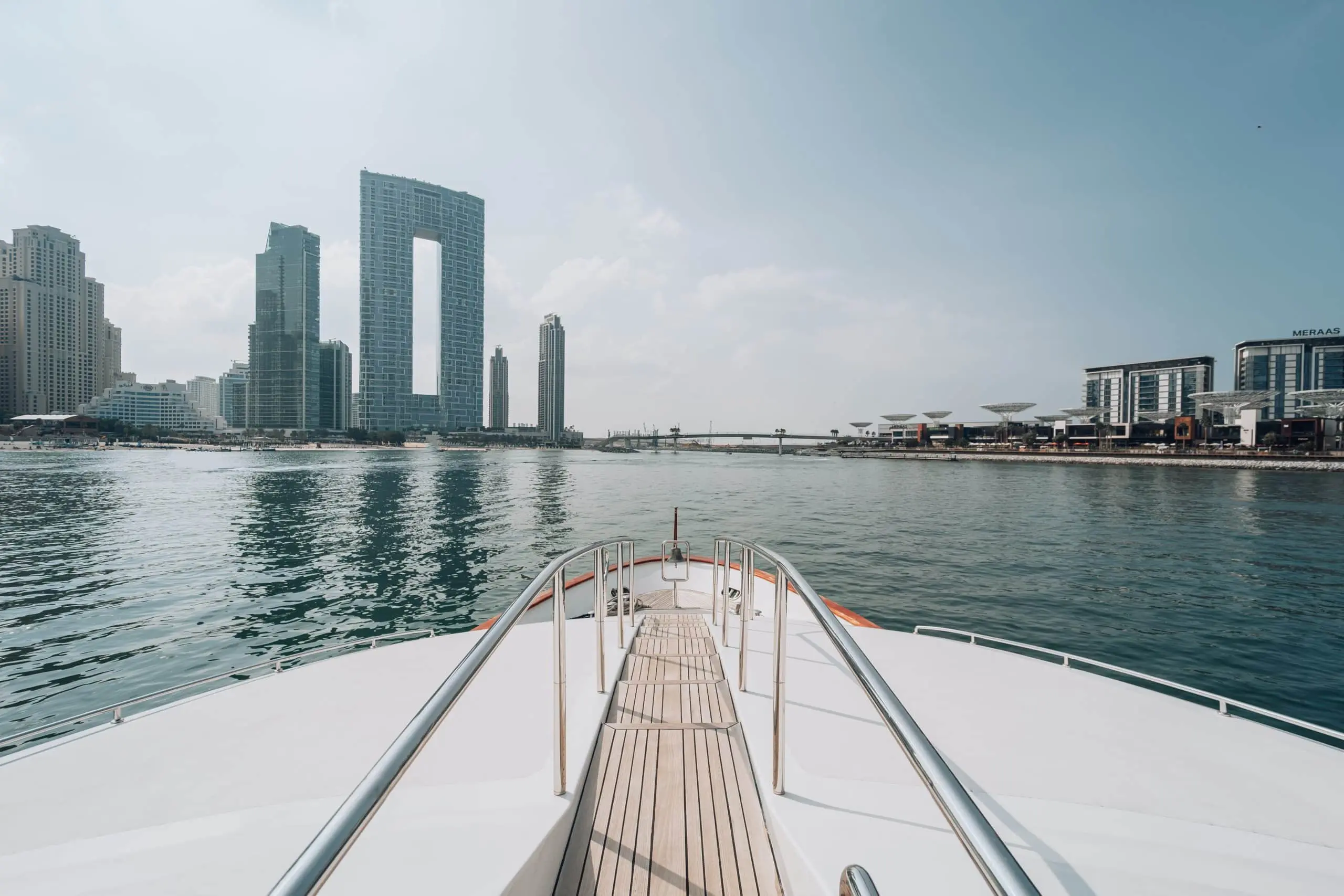 View from the bow of a luxury yacht looking forward over calm water toward a modern waterfront skyline, featuring a distinctive high-rise building with a large rectangular opening.