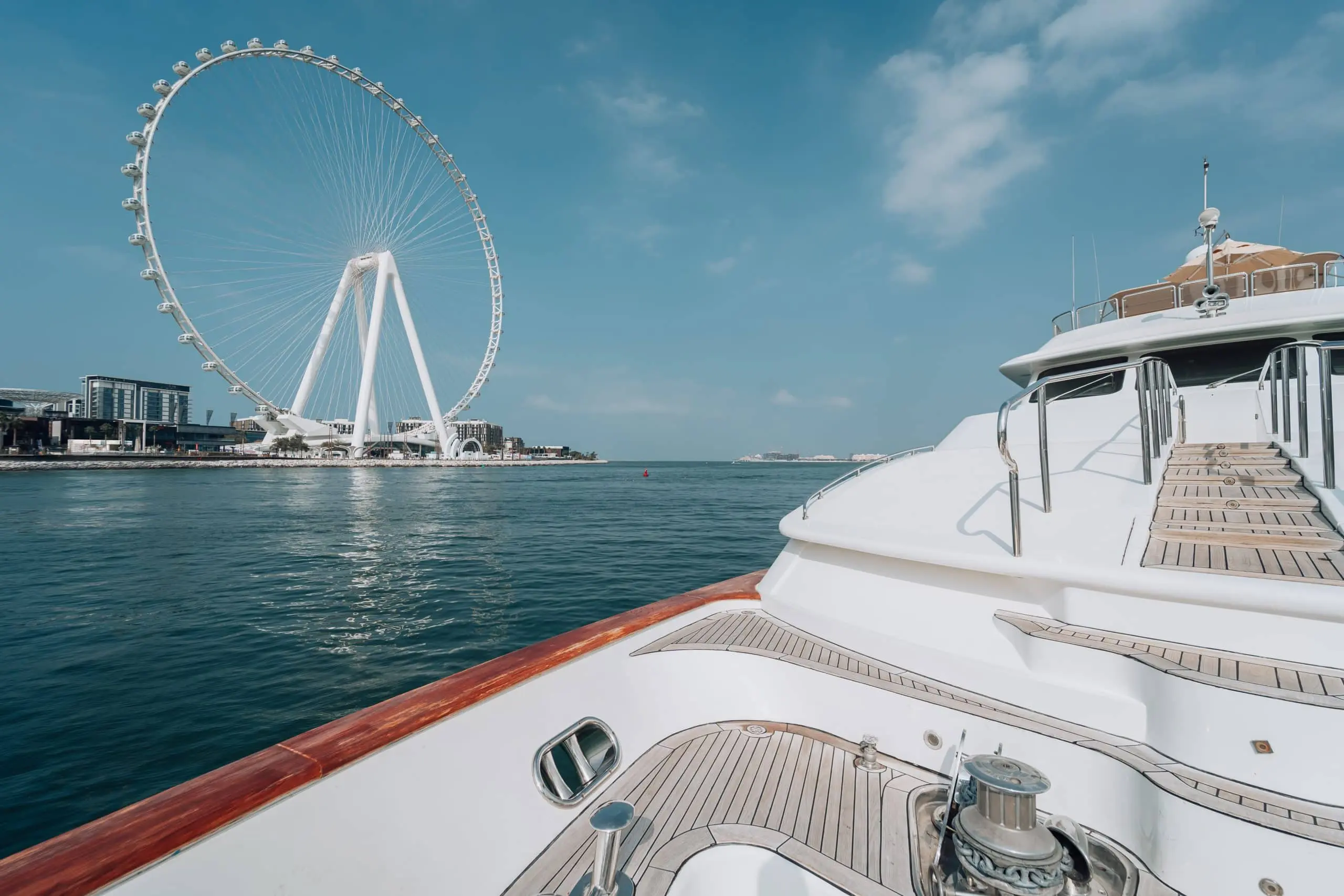 View from the bow of a luxury yacht facing calm water and a large waterfront observation wheel in the distance.