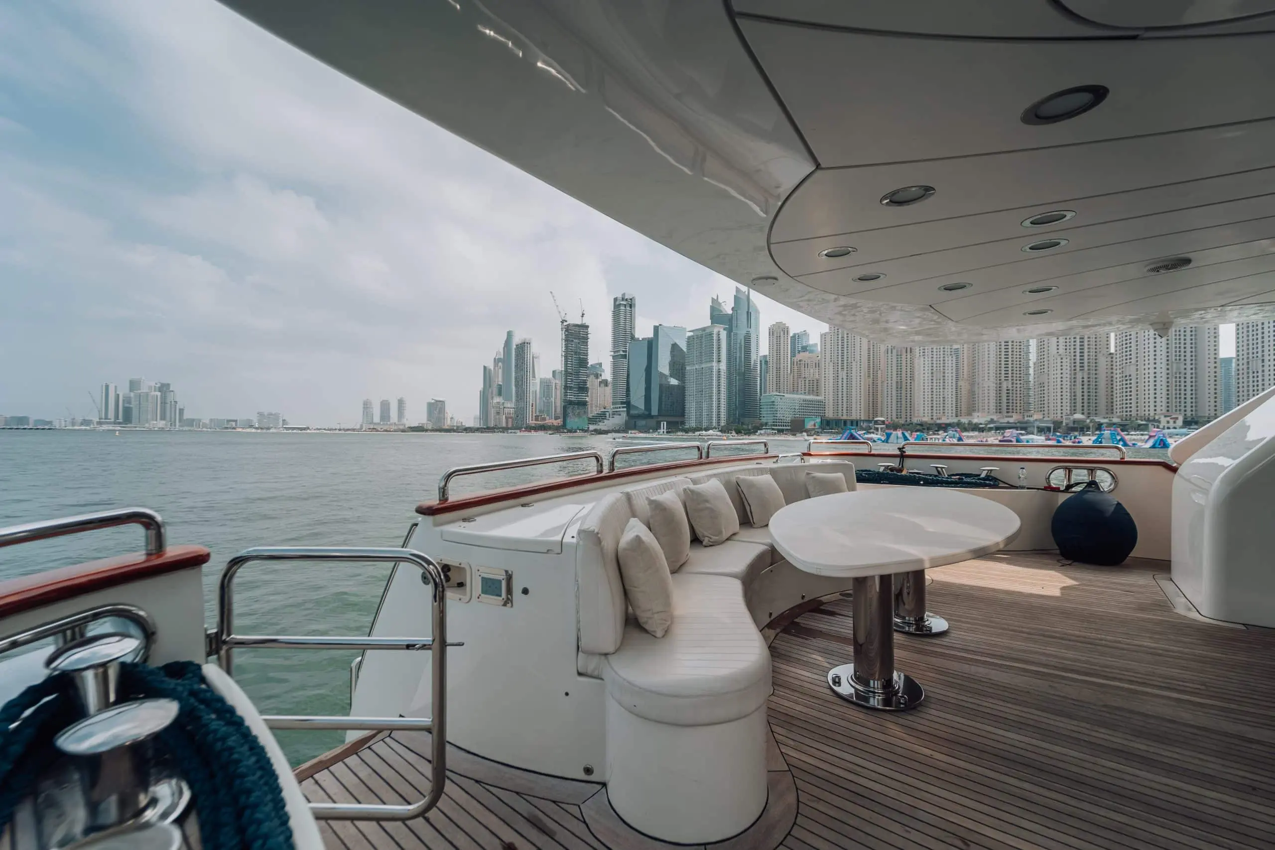 Curved built-in sofa and round dining table on the shaded rear deck of a luxury yacht, overlooking calm water and a modern city skyline across the marina.