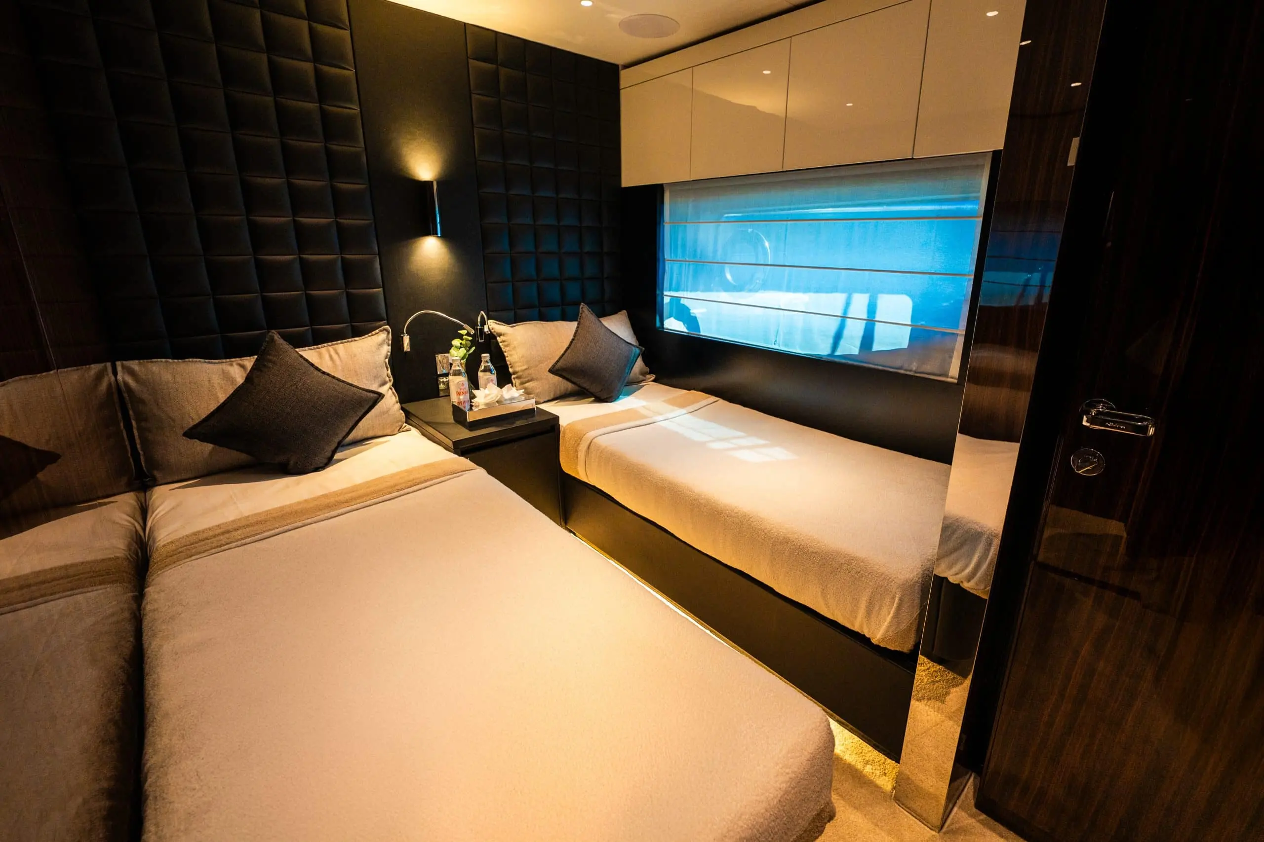 Luxury yacht twin cabin with padded walls, twin beds, and blue-lit porthole window