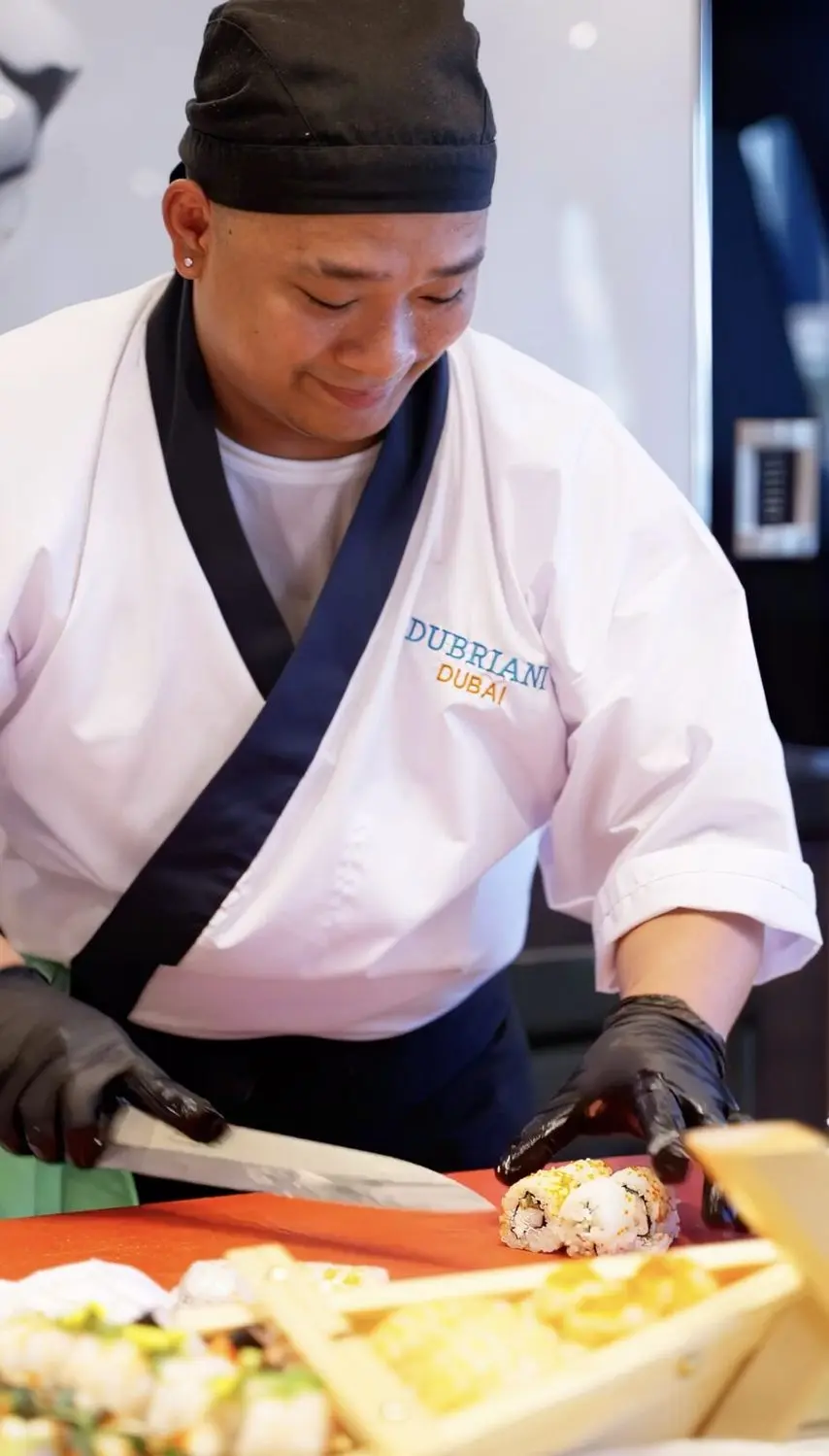 Chef in a Dubriani Dubai jacket slicing a sushi roll on a wooden board during onboard catering
