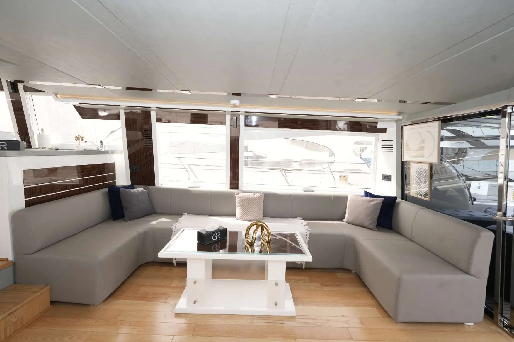 Spacious salon interior on Belle 75 yacht featuring U-shaped grey leather sofa, glass coffee table, and panoramic windows with marina views.