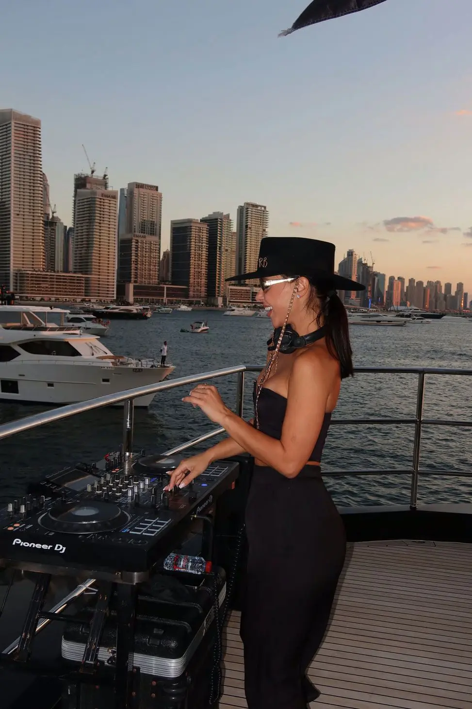 Female DJ performing live on luxury yacht at sunset with Dubai skyline in background