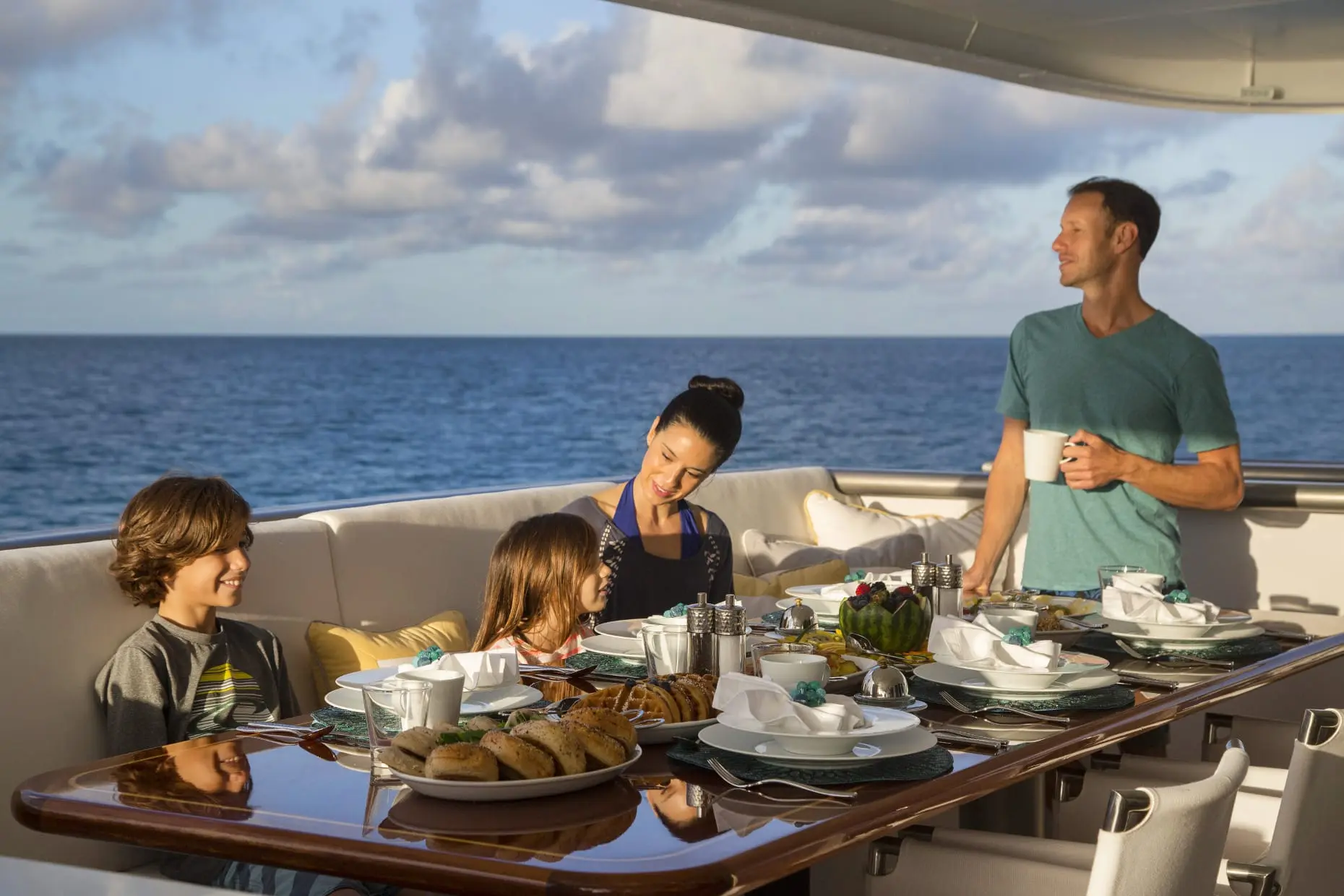 Family sharing a meal at a laid table on a yacht's aft deck with open sea behind
