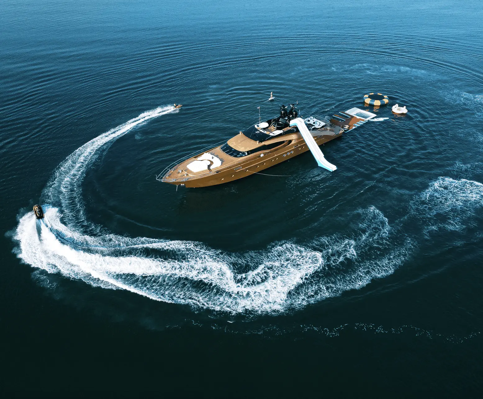 Luxury royal yacht anchored in deep blue water with a waterslide, floating platforms, and jet skis creating circular wake patterns nearby.