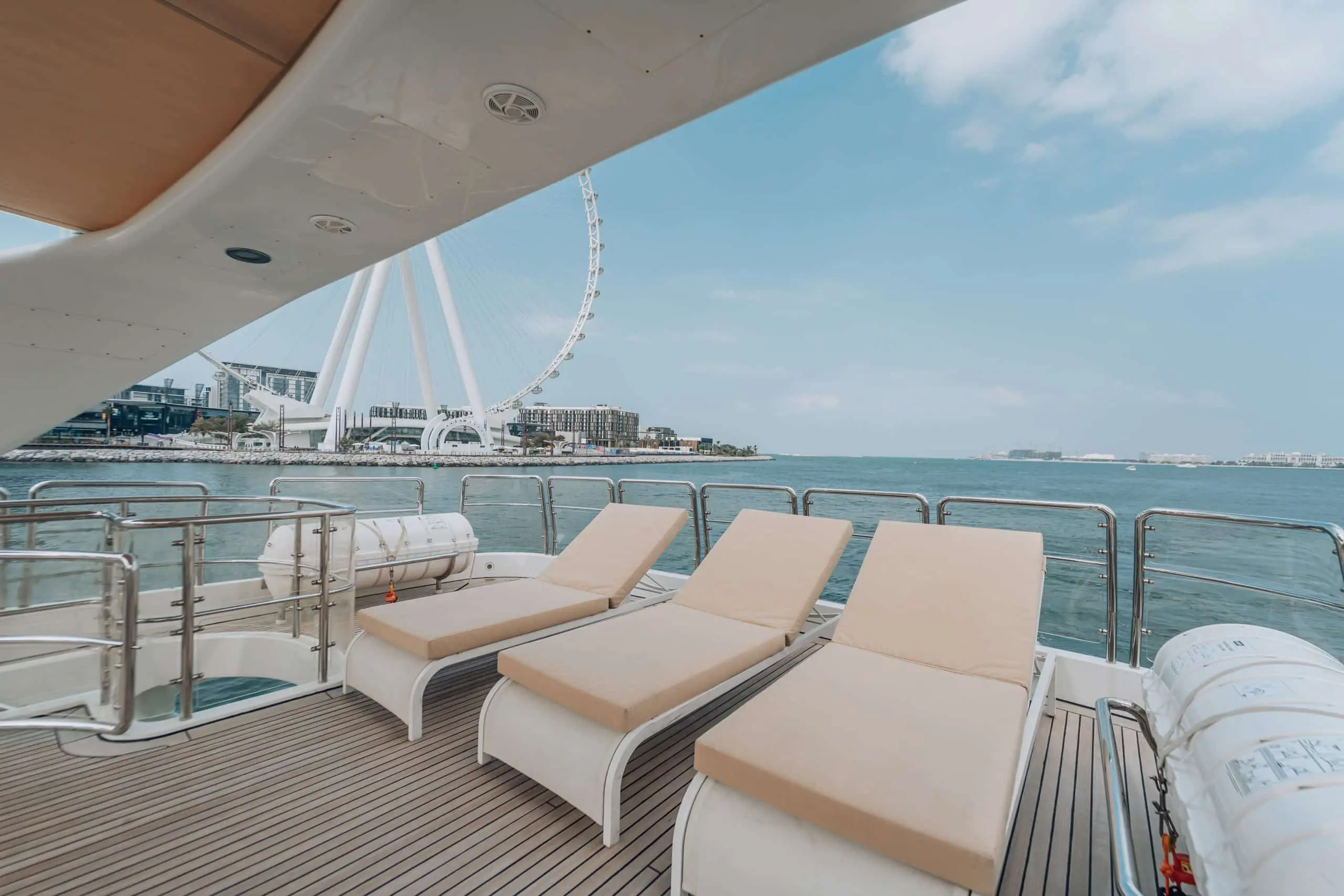 Yacht deck with sun loungers near waterfront Ferris wheel.