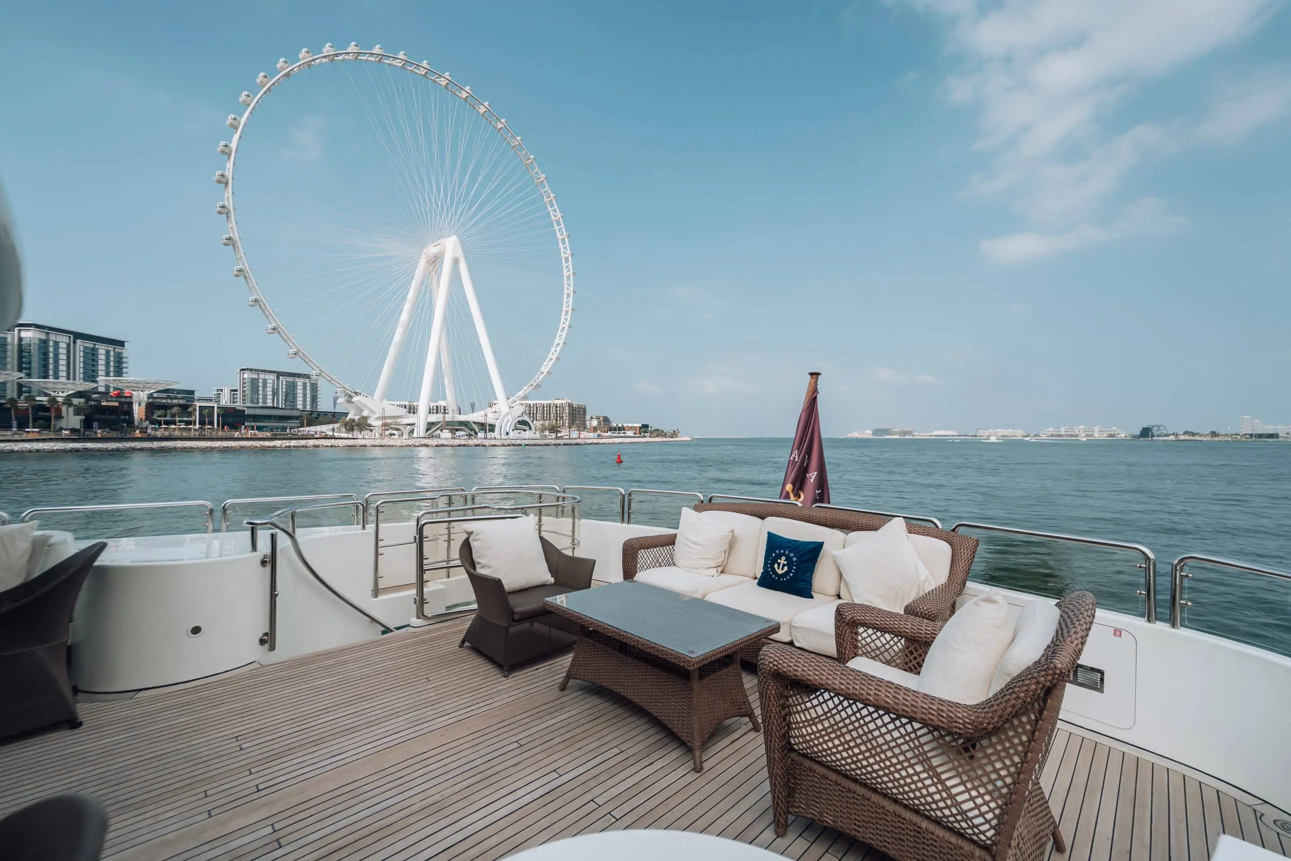 Lounge seating on the open deck of a luxury yacht, with wicker chairs and a sofa facing calm water and a large observation wheel and modern waterfront buildings in the background.