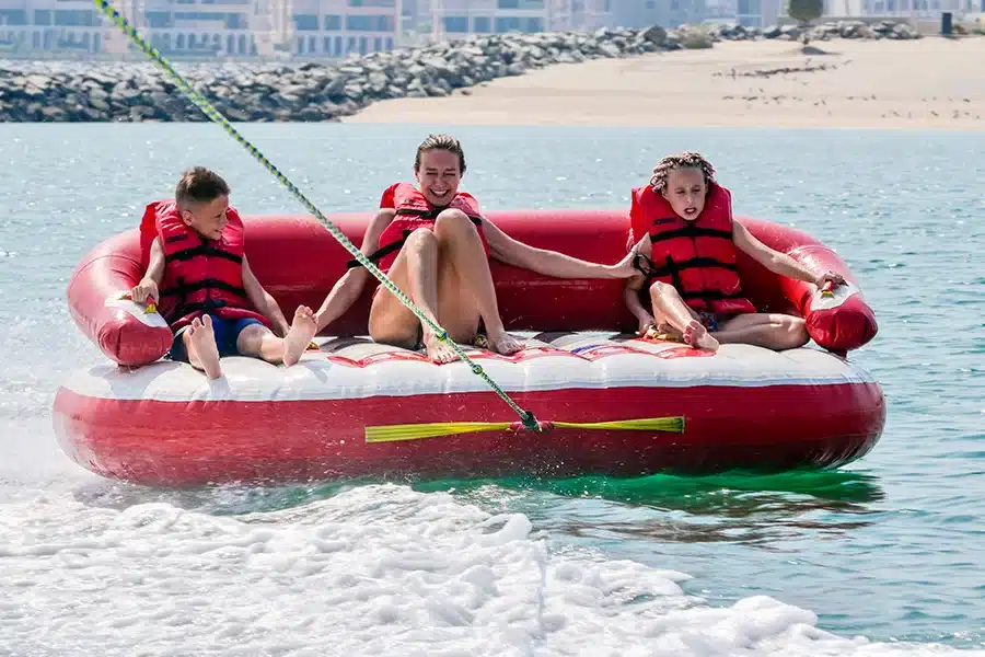 Three riders in red life jackets on an inflatable donut tube towed across turquoise water near the beach