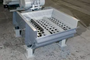 Industrial vibrating screen with perforated metal tray on a steel frame and motor above, used to sort materials on a factory floor.