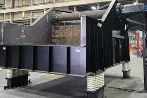 Large industrial machine with a rotating roller, metal housing, and coil-spring supports on a factory floor.