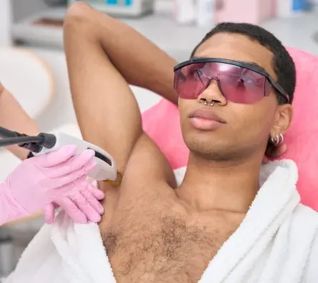 Young man undergoing laser epilation procedure in beauty clinic Male in safety goggles lying supine during armpit hair removal conducted by cosmetologist in nitrile gloves