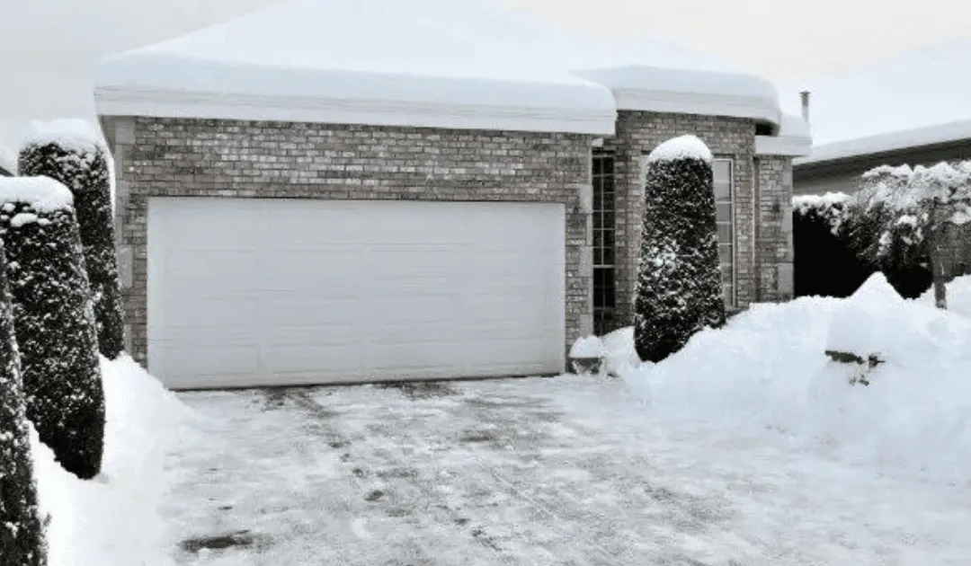 Snow-covered brick house with a garage and trimmed bushes in winter.