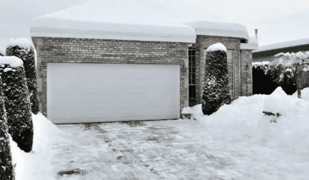 Snow-covered brick house with a garage and trimmed bushes in winter.