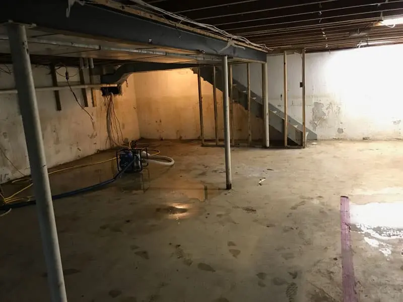 Basement interior showing damp concrete floor, exposed pipes, and stairs, emphasizing the need for waterproofing solutions to prevent water intrusion.