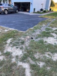 Drainage pipe outlet in yard beside asphalt driveway, with grass and dirt surrounding, illustrating landscape grading for water runoff management.