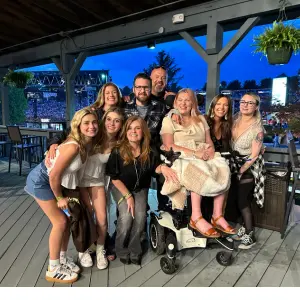 Group of people posing together, including a woman in a mobility scooter, at an outdoor event, showcasing community support and solidarity for ALS advocacy.