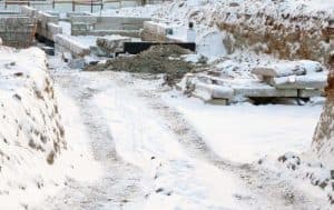 Construction site in winter with snow-covered ground, exposed foundation blocks, and gravel, illustrating cold-weather conditions affecting basement waterproofing efforts.