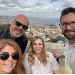Group of four smiling individuals posing for a selfie with a scenic city view in the background, highlighting camaraderie and support for community initiatives.