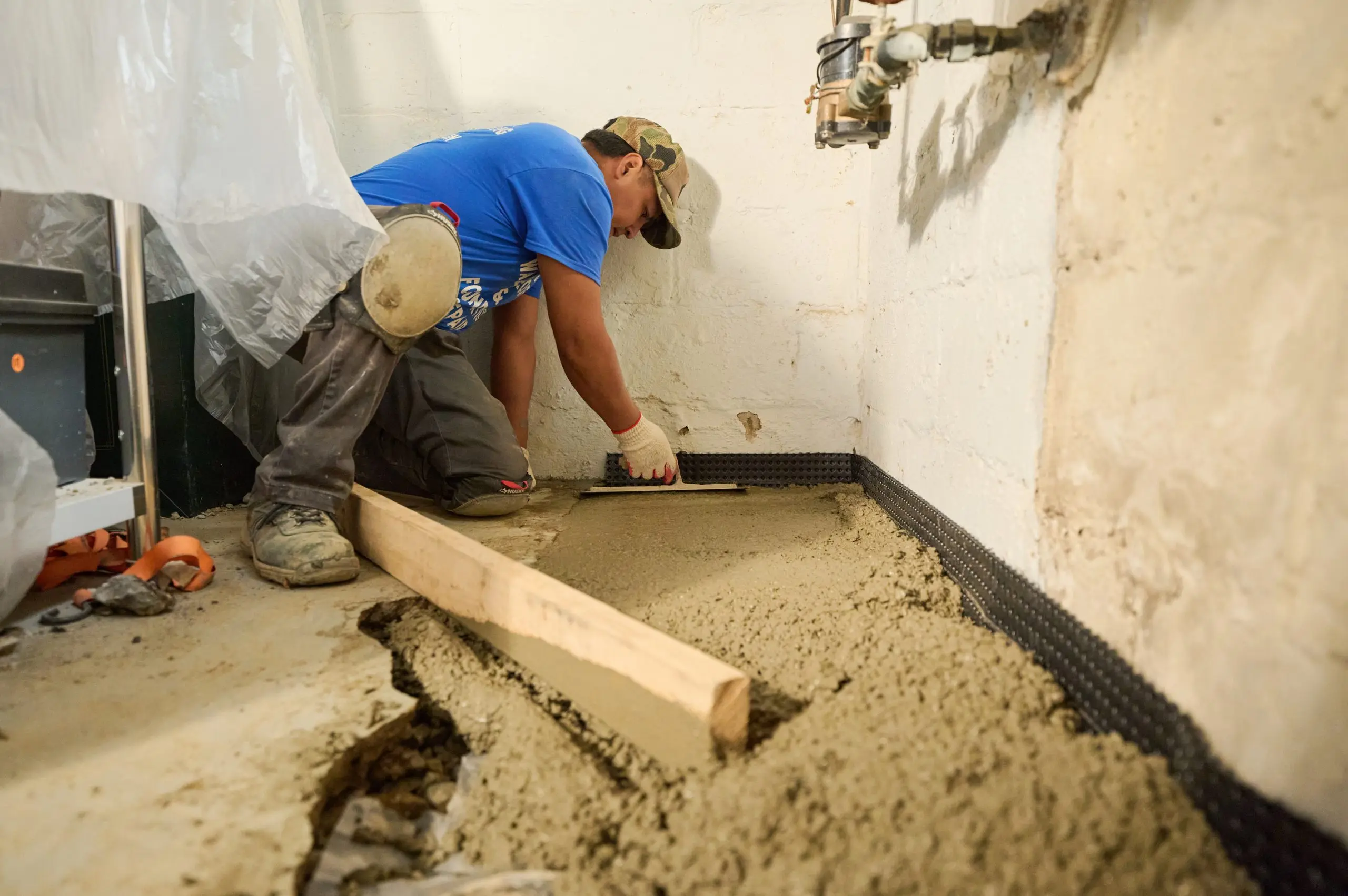 Worker in cap and blue shirt smoothing wet concrete on a basement floor, surrounded by construction tools and pipes, conveying focus and diligence.