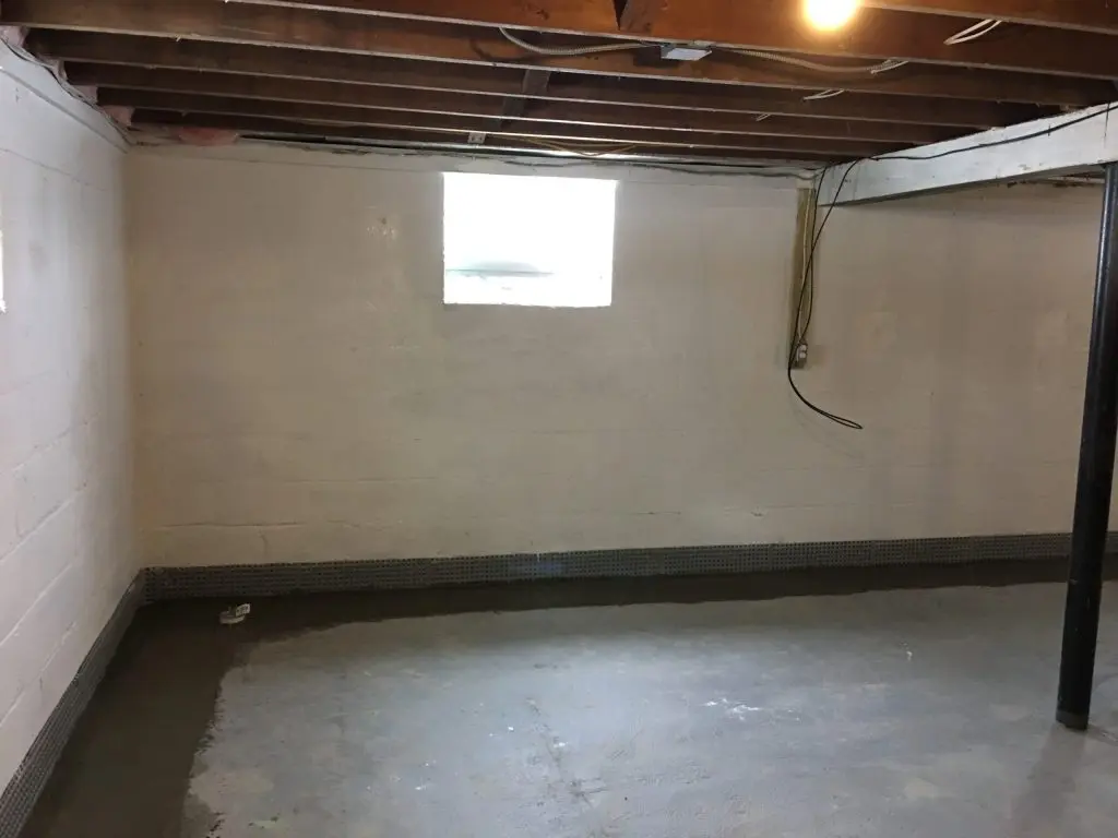 Basement interior with newly installed drainage system along the perimeter, showcasing dry concrete floor and window, emphasizing waterproofing solutions for water intrusion prevention.