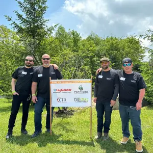 Four men in black Highlander Waterproofing t-shirts standing beside a sponsorship sign at the 8th Annual Sporting Clay Shoot & Fundraiser, promoting community support and initiatives against human trafficking.