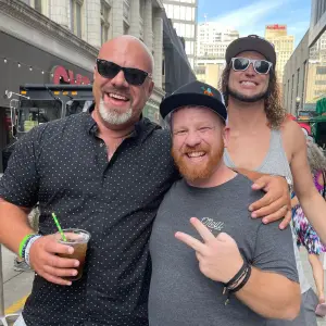 Three men smiling and posing together at a community event, celebrating culture and togetherness, with one holding a drink, in an outdoor setting reflecting the vibrant atmosphere of Rock Reggae Relief.