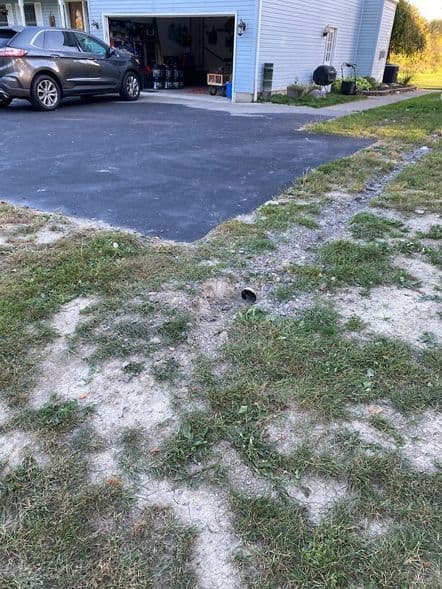 Drainage pipe outlet in grassy area near garage, highlighting potential water flow direction and grading concerns for effective drainage management.
